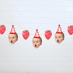May include: A festive banner featuring a string with alternating images of strawberries and baby faces wearing party hats. The hats are pink with white polka dots and the number "1". The strawberries are red.