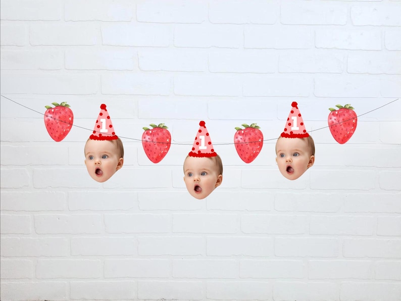 May include: A festive banner featuring a string with alternating images of strawberries and baby faces wearing party hats. The hats are pink with white polka dots and the number "1". The strawberries are red.