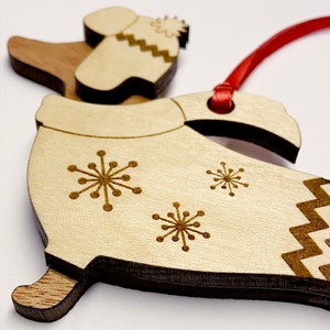 K&ouml;nnte beinhalten: Weihnachtsbaumschmuck aus Holz in Form einer Person, die einen Winterhut und einen Mantel mit Schneeflockenmotiven tr&auml;gt. Der Schmuck ist an einem roten Band befestigt.