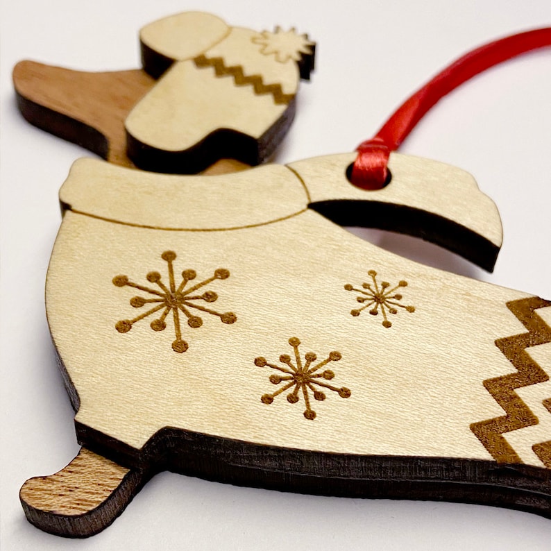 K&ouml;nnte beinhalten: Weihnachtsbaumschmuck aus Holz in Form einer Person, die einen Winterhut und einen Mantel mit Schneeflockenmotiven tr&auml;gt. Der Schmuck ist an einem roten Band befestigt.