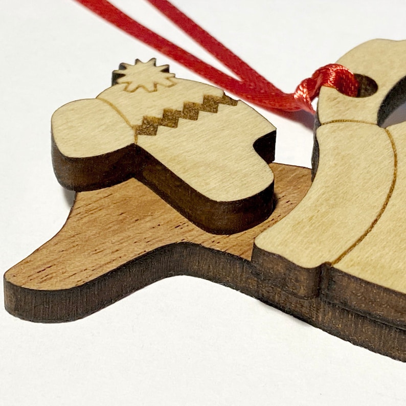 K&ouml;nnte beinhalten: Ein h&ouml;lzerner Weihnachtsbaumschmuck mit einer Glocke und einem Hut mit Schneeflockenmuster. Der Anh&auml;nger ist an einem roten Band befestigt.