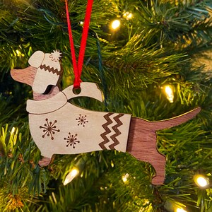 K&ouml;nnte beinhalten: Weihnachtsbaumschmuck aus Holz in Form eines Dackels, der eine wei&szlig;e Strickm&uuml;tze und einen Pullover mit Schneeflockenmuster tr&auml;gt. Der Schmuck h&auml;ngt an einem Weihnachtsbaum.