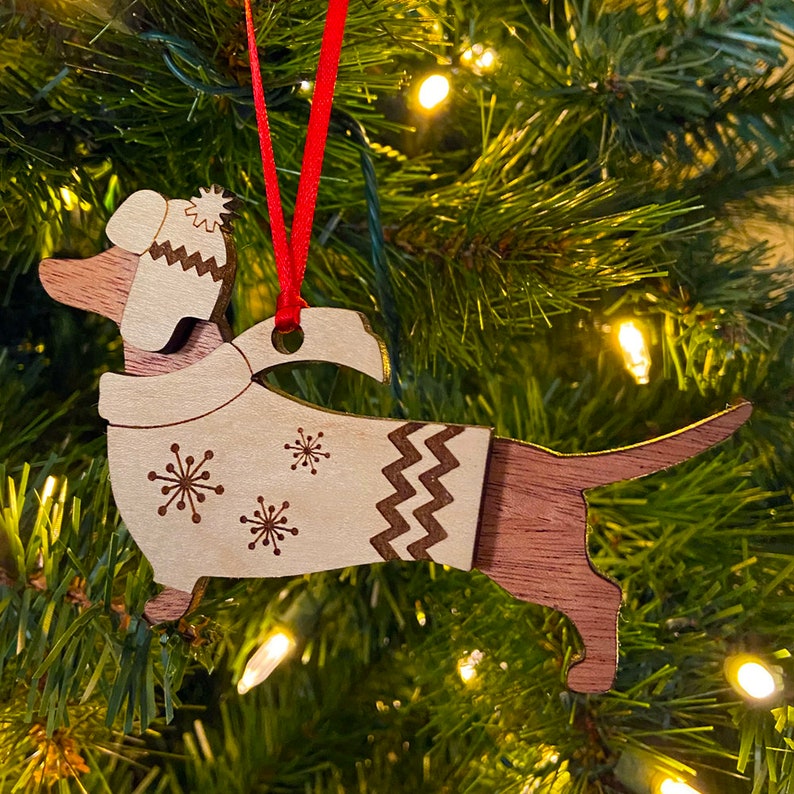 K&ouml;nnte beinhalten: Weihnachtsbaumschmuck aus Holz in Form eines Dackels, der eine wei&szlig;e Strickm&uuml;tze und einen Pullover mit Schneeflockenmuster tr&auml;gt. Der Schmuck h&auml;ngt an einem Weihnachtsbaum.