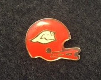 Op de afbeelding: Een rood-witte voetbalhelmspeld. De helm is rood met een wit gezichtsmasker en een witte streep aan de zijkant.