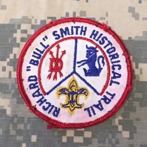 May include: A red, white, and blue embroidered patch with the text "Richard "Bull" Smith Historical Trail". The patch features a red, white, and blue design with a fleur-de-lis in the center.