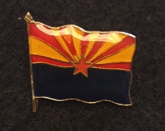 Op de afbeelding: Een gouden speld met de vlag van de staat Arizona. De vlag is rood, geel en blauw met een rode ster in het midden.