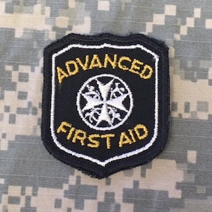 May include: Black and white patch with a white cross in the center and the text "ADVANCED FIRST AID" in yellow letters.