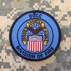 May include: A blue and yellow round patch with the text "DSCC LOGISTICS AGENCY DIVISION OF FIRE" and a stylized eagle with a shield.