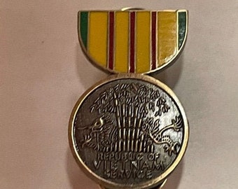 Op de afbeelding: Een Vietnam Service medaille-speld met een goudkleurige rand. De speld heeft een geel en rood gestreept lint boven een cirkelvormig embleem met de woorden "REPUBLIC OF VIETNAM SERVICE". Het embleem heeft een bamboebosontwerp.