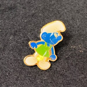 May include: A gold-colored pin featuring a cartoon Smurf character. The Smurf is wearing a green and blue outfit and has a white hat. The pin is a collectible item.