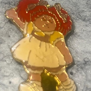 May include: A gold-toned metal enamel pin featuring a doll with bright red yarn-style hair, wearing a cream-colored dress with yellow accents and white shoes. The doll is depicted with one arm raised in a waving gesture, finished with a polished gold outline.