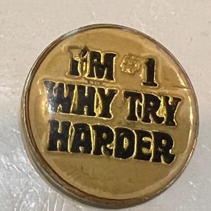 May include: A round, gold-colored pin with the text "I'M #1 WHY TRY HARDER" in black lettering. The pin has a slightly worn appearance, suggesting it may be vintage or pre-owned. The pin is a motivational or humorous item.