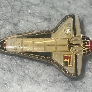 May include: A metal enamel pin shaped like a space shuttle, featuring a white body with black and gold trim. The text USA is printed on the wing, and a small American flag graphic is visible. The nose cone has red accents, and the tail has black details.