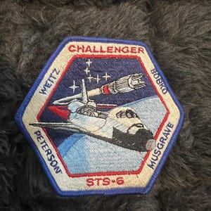 May include: A blue and white embroidered patch with the text "CHALLENGER STS-6" and the names "WETTZ, BOBKO, PETERSON, MUSGRAVE" around the perimeter. The patch features a space shuttle in flight with a red, white, and blue color scheme.