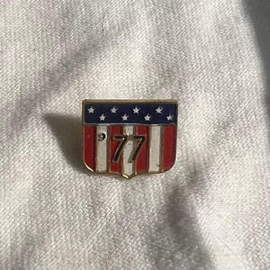 May include: A gold-colored pin with a red, white, and blue American flag design. The number 77 is printed in black on the flag.