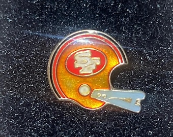 Op de afbeelding: Een gouden, rode en witte voetbalhelmspeld. De speld heeft het logo van het team in het midden van de helm. De helm heeft een gouden gezichtsmasker en een rode streep.