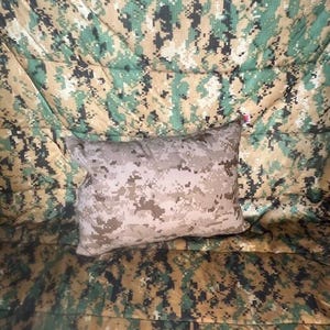 May include: A tan and brown camouflage pillow on a green, tan, and brown camouflage background.