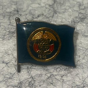 May include: A rectangular metal lapel pin shaped like a waving flag with a dark blue enamel finish. Centered on the flag is a gold circular emblem featuring a stylized eagle above a shield with red and gold details and the year 1896 printed below.
