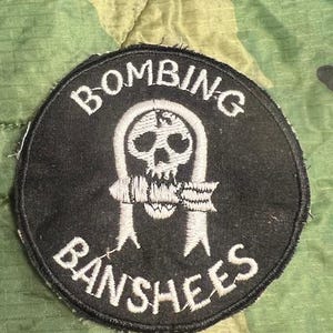 May include: A black and white embroidered patch with a skull and crossbones design. The text "Bombing Banshees" is stitched around the skull.