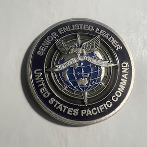 May include: A silver challenge coin with a blue and silver design. The coin features a globe with a compass rose, an eagle, and the text "Senior Enlisted Leader United States Pacific Command".
