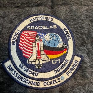 May include: A blue and white embroidered patch with the text "SPACELAB" and the names of the astronauts: "HARTFIELD", "BUCHLI", "MESSERSCHMID", "BLUFORD", "OCKELS", "DUNBAR", "FURRER", and "NAGEL". The patch also features a space shuttle, a globe, and the number "01".