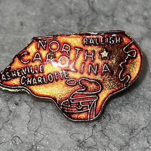 May include: A North Carolina state-shaped pin with a map design. The pin features the state's name and the cities of Asheville, Charlotte, and Raleigh. The pin has a gold border and a red, orange, and yellow color scheme.