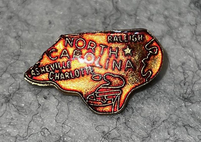 May include: A North Carolina state-shaped pin with a map design. The pin features the state's name and the cities of Asheville, Charlotte, and Raleigh. The pin has a gold border and a red, orange, and yellow color scheme.