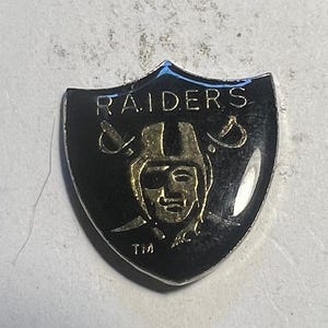 May include: A black and silver Raiders shield-shaped pin. The pin features the word "RAIDERS" in silver, a gold helmeted football player, and two crossed swords.
