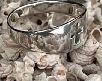 Op de afbeelding: Een zilveren ring met een gehammerde afwerking en een kruisontwerp. De ring ligt op een bed van witte schelpen.