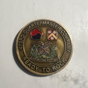 May include: A gold-colored coin with the text "473rd Quartermaster Company" and "Ready to Rock" around the edge. The center features a military insignia with a red, white, and blue design and the text "Supporting Victory".