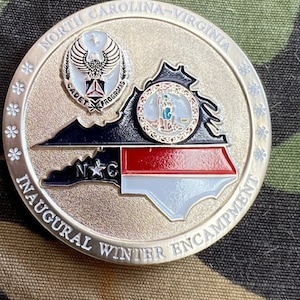 May include: A silver coin with a gold outline featuring a map of North Carolina and Virginia with the text "North Carolina-Virginia Inaugural Winter Encampment" around the edge. The coin also features a seal with the text "Cadet Programs" and a star.