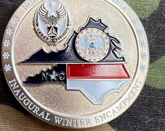 Op de afbeelding: Een zilveren munt met een gouden rand met een kaart van North Carolina en Virginia met de tekst "North Carolina-Virginia Inaugural Winter Encampment" rond de rand. De munt heeft ook een zegel met de tekst "Cadet Programs" en een ster.