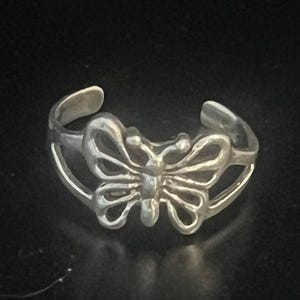 May include: A silver-colored ring with a butterfly design. The butterfly has detailed wing patterns and is the focal point of the ring. The ring is set against a dark background.