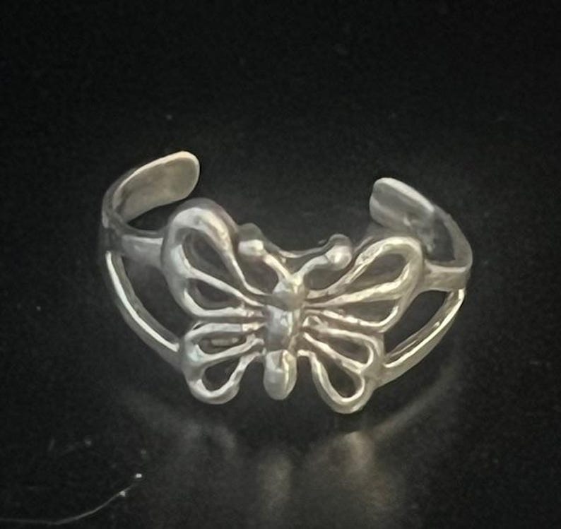 May include: A silver-colored ring with a butterfly design. The butterfly has detailed wing patterns and is the focal point of the ring. The ring is set against a dark background.