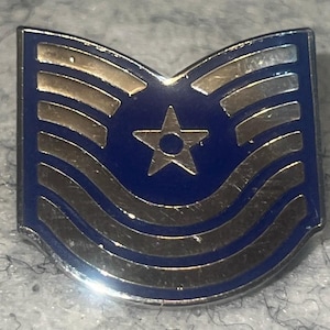 May include: A metal pin featuring a blue enamel background with silver-toned stripes and a central five-pointed star. The design is shaped like a chevron with a curved top edge and a rounded bottom edge, reflecting a military-style rank insignia.