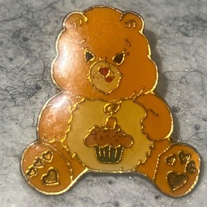 May include: A gold-toned metal enamel pin shaped like an orange bear with a light yellow belly patch. The bear features a brown cupcake graphic with a single lit candle on its stomach, and three small heart-shaped indentations on each foot.
