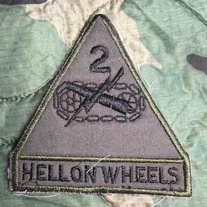 May include: A green triangular patch with a black embroidered image of a cannon and the number "2" above it. The text "HELL ON WHEELS" is embroidered in black at the bottom of the patch.