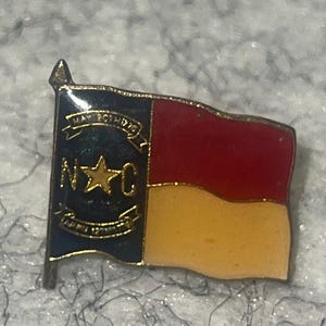 May include: A metal lapel pin shaped like the North Carolina state flag. The design features a blue vertical bar with a gold star, the letters N and C, and two gold banners with the text MAY 20TH 1775 and APR 12TH 1776. The right side has red and white horizontal stripes.