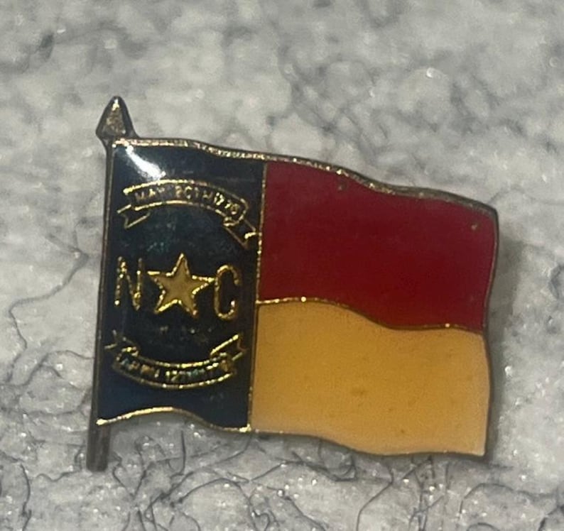 May include: A metal lapel pin shaped like the North Carolina state flag. The design features a blue vertical bar with a gold star, the letters N and C, and two gold banners with the text MAY 20TH 1775 and APR 12TH 1776. The right side has red and white horizontal stripes.