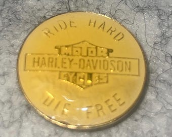 Op de afbeelding: Een ronde, goudkleurige Harley-Davidson-speld met de woorden "RIDE HARD DIE FREE" langs de rand. Het Harley-Davidson-logo staat in het midden.