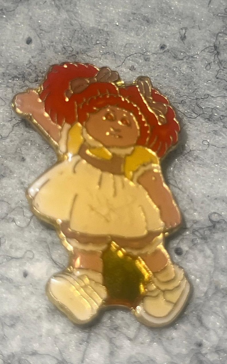 May include: A gold-toned metal enamel pin featuring a doll with bright red yarn-style hair, wearing a cream-colored dress with yellow accents and white shoes. The doll is depicted with one arm raised in a waving gesture, finished with a polished gold outline.