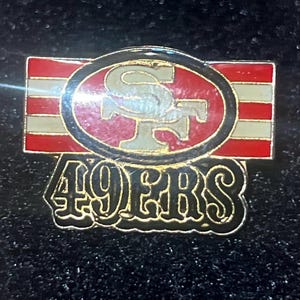 May include: A metal pin featuring the San Francisco 49ers logo. The pin has a red, white, and gold color scheme. The team's logo is in the center, with the words "49ERS" below it.