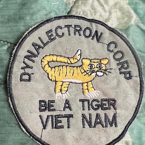 May include: A green, brown, and beige fabric patch with black embroidered text that reads "Dynalectro Corp." and "Be a Tiger Viet Nam". A yellow embroidered tiger is in the center of the patch.