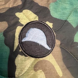 May include: A black and white military patch with a blue helmet design. The patch is circular and has a white border.