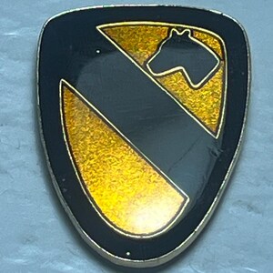 May include: A black and gold shield-shaped pin with a horse head silhouette. The pin features a black stripe and gold accents, with a gold border. The design is a military insignia.