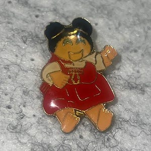 May include: A gold-toned metal enamel pin featuring a cartoon character with dark hair in two buns, wearing a red dress with a white collar and a small gold detail on the chest. The character has a tan complexion, green eyes, and is depicted in a seated position.