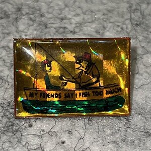 May include: A rectangular pin with a gold holographic background featuring a graphic of two figures in a boat fishing. The boat has the text "MY FRIENDS SAY I FISH TOO MUCH" printed on the side. The water below the boat is dark green.