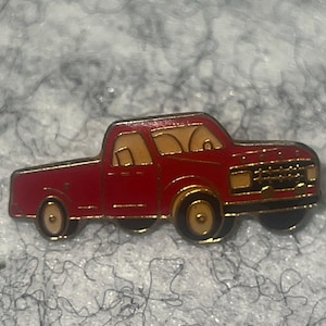 May include: A red pickup truck pin with gold-colored accents. The pin features a detailed design of a classic truck, including the cab, bed, and wheels. The pin is likely made of metal and has a glossy finish.