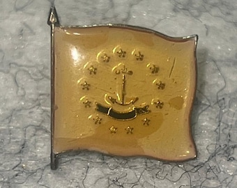 Op de afbeelding: Een rechthoekige metalen reversspeld in de vorm van een vlag met een lichtbruine emaille afwerking. Het ontwerp toont een centraal goudkleurig anker omringd door een cirkel van dertien kleine gouden sterren en een horizontale banner onderaan.