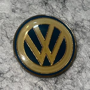 May include: A round, enamel pin featuring the Volkswagen logo. The pin has a blue and cream-colored design with a gold outline. The VW logo is in the center, with a blue background and cream-colored border.